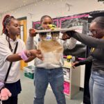 "Three students examining a plant's roots in a classroom setting."